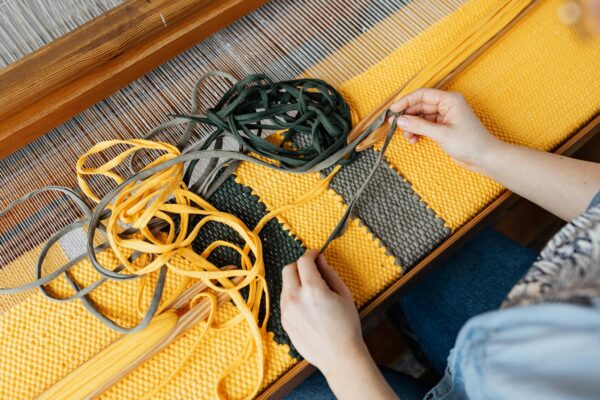 Top view of crop anonymous female employee working on wooden weaving loom machine with stretched durable colorful threads in workshop