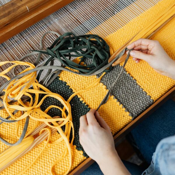 Top view of crop anonymous female employee working on wooden weaving loom machine with stretched durable colorful threads in workshop